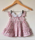 A baby girl's light pink tiered dress with an all-over pink and purple floral print, featuring a contrasting white Peter Pan collar and ruffled flutter sleeves