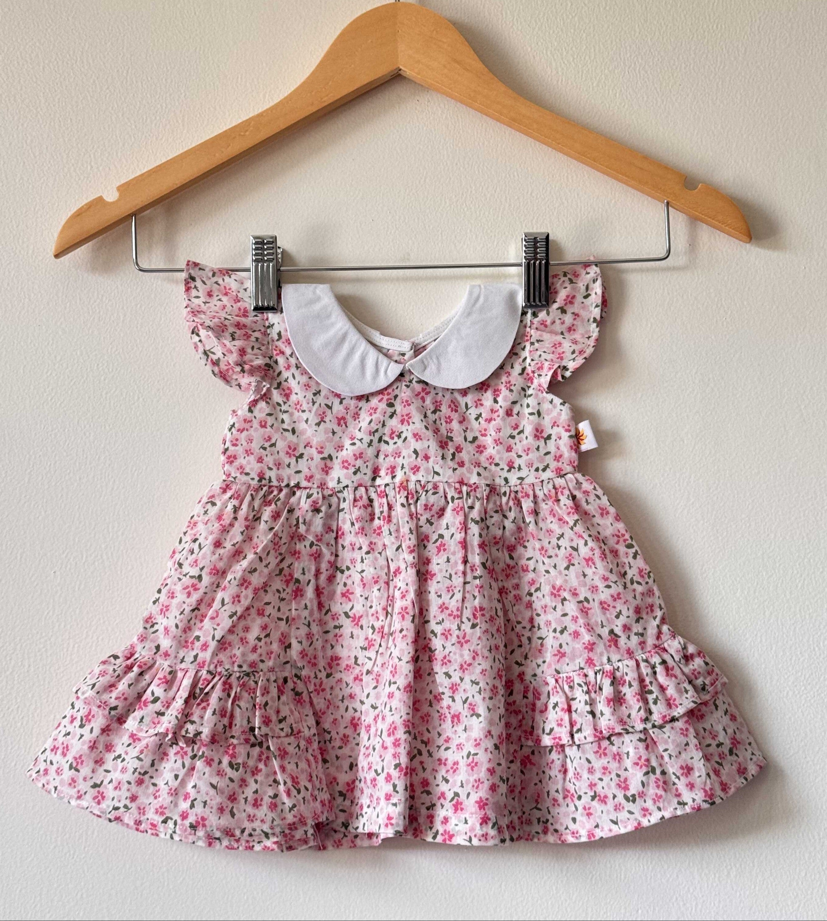A baby girl's light pink tiered dress with an all-over pink and purple floral print, featuring a contrasting white Peter Pan collar and ruffled flutter sleeves