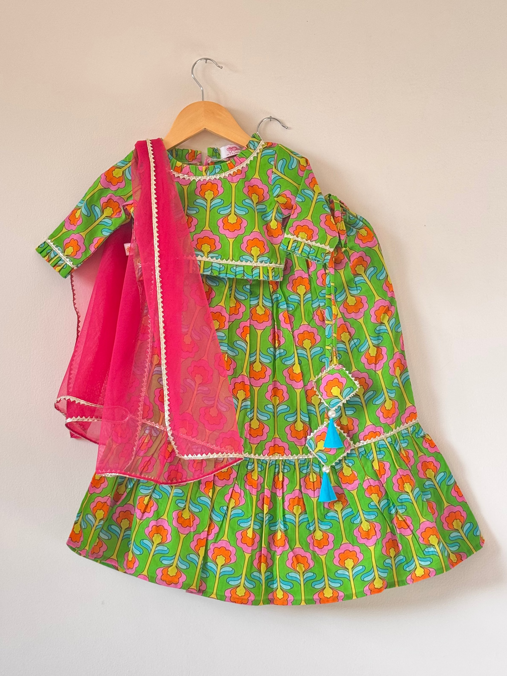 A children's vibrant green traditional lehenga choli set. The outfit features a high-definition digital floral print in pink and yellow with soft gold lace trims on the round neck, cuffs, and hem. It includes a long-sleeve choli top, a tiered flared lehenga skirt with a light blue tassel accent, and a draped translucent hot pink net dupatta.