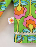 Close-up of a kids' long-sleeve choli top featuring a vibrant green fabric with large, stylized retro floral patterns in orange, pink, yellow, and blue. The design highlights a small, white woven "Cheera Meera" brand tag with an orange flower logo sewn into the side seam near the cuff, which is finished with delicate, soft gold pleated lace trim.