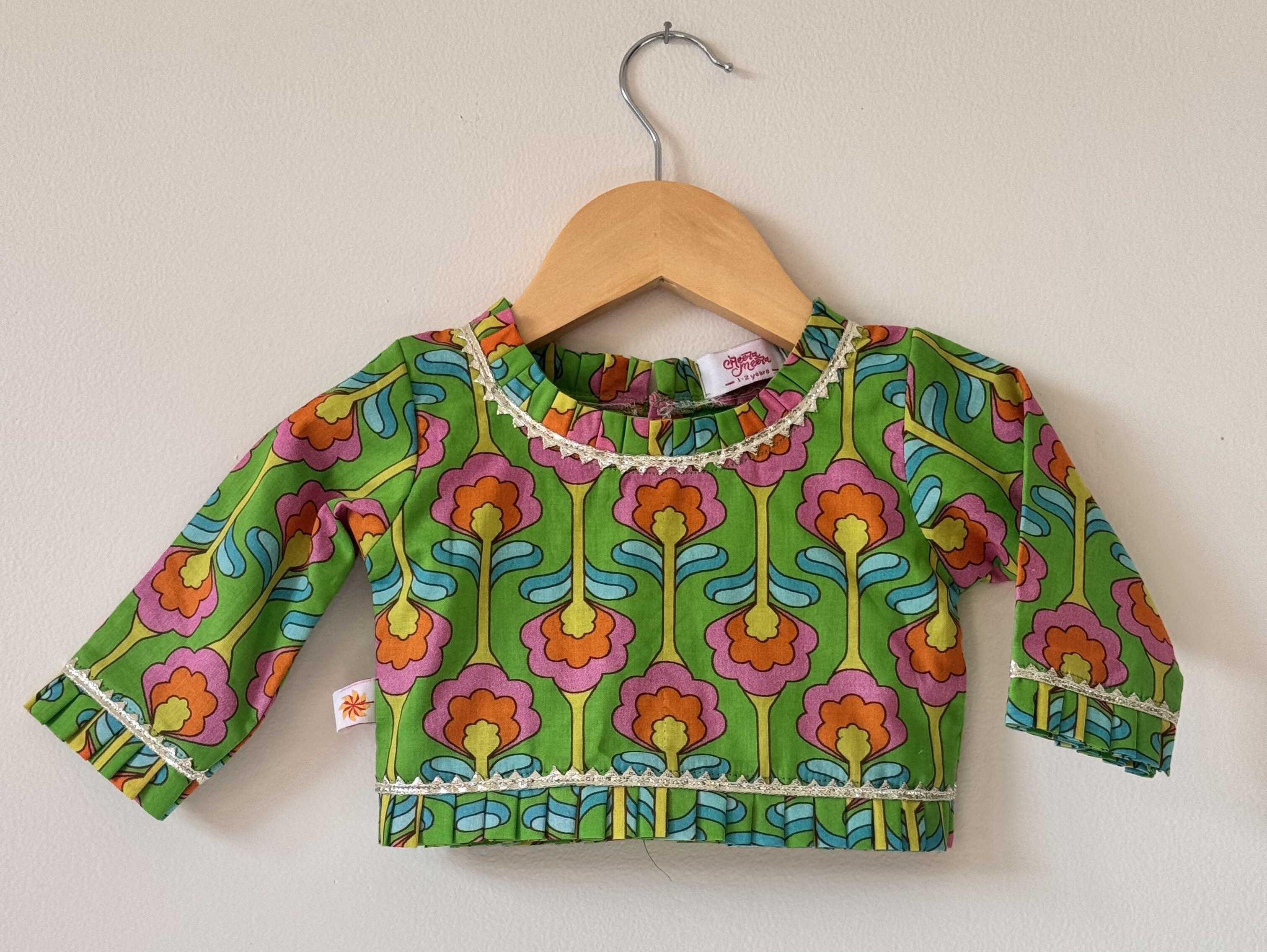 Close-up of a kids' green long-sleeve choli top. The top features a vibrant digital floral pattern in pink, orange, yellow, and green, complete with soft gold pleated lace trim detailing along the round neckline, sleeve cuffs, and bottom hem.