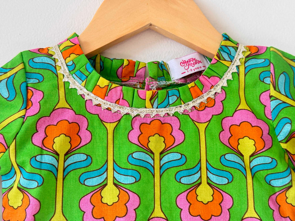 Close-up of the neckline on a kids' green choli top. It features a vibrant, symmetrical digital print with large pink and orange floral motifs, a round neckline lined with delicate white pleated lace trim, and a small woven "Cheera Meera" brand tag.