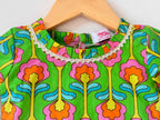 Close-up of the neckline on a kids' green choli top. It features a vibrant, symmetrical digital print with large pink and orange floral motifs, a round neckline lined with delicate white pleated lace trim, and a small woven "Cheera Meera" brand tag.