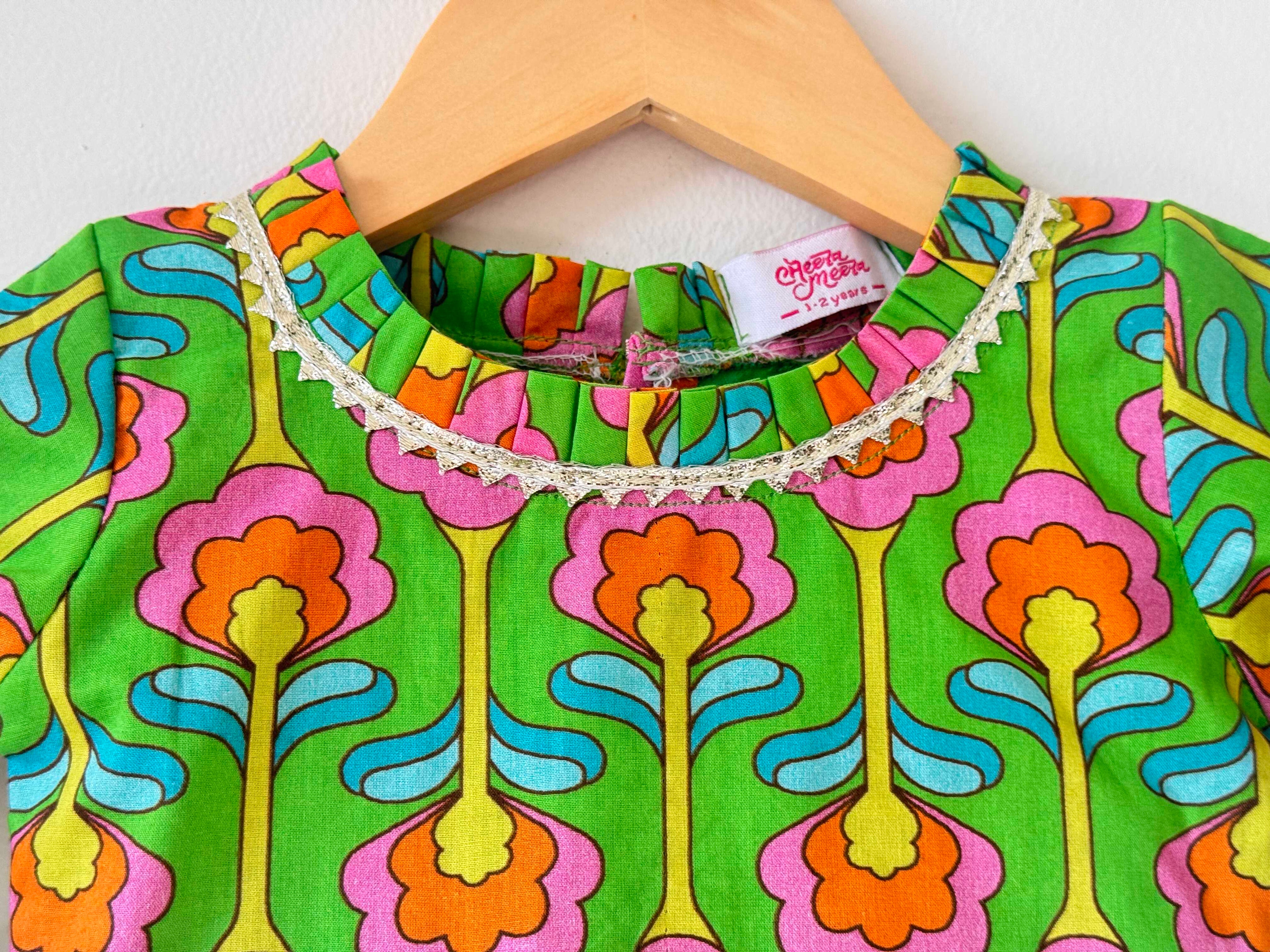 Close-up of the neckline on a kids' green choli top. It features a vibrant, symmetrical digital print with large pink and orange floral motifs, a round neckline lined with delicate white pleated lace trim, and a small woven "Cheera Meera" brand tag.