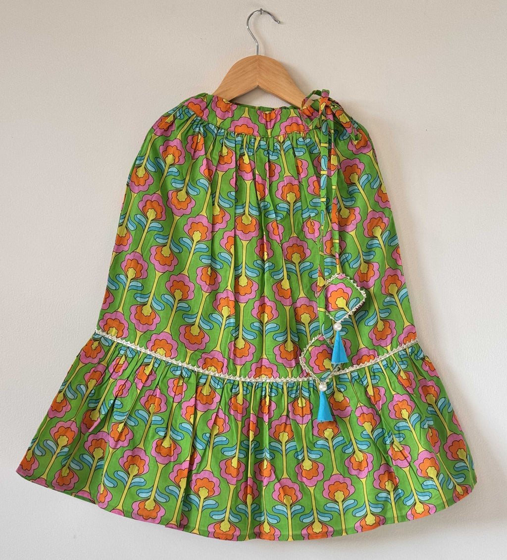 A kids' vibrant green cotton lehenga skirt hanging on a wooden hanger against a plain background. The skirt features an all-over digital floral print in pink, orange, and yellow, a tiered flared hemline trimmed with delicate white pleated lace, and two light blue tassel ties at the waist.