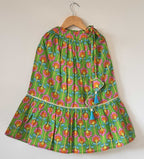A kids' vibrant green cotton lehenga skirt hanging on a wooden hanger against a plain background. The skirt features an all-over digital floral print in pink, orange, and yellow, a tiered flared hemline trimmed with delicate white pleated lace, and two light blue tassel ties at the waist.