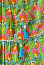 Close-up of the waistband area on a kids' green cotton lehenga skirt. The image showcases two bright blue fabric tassels with small white pearl bead accents, hanging from a thin white cord. The background features a vibrant digital floral print in shades of orange, pink, yellow, and blue, split by a delicate soft gold lace trim line.