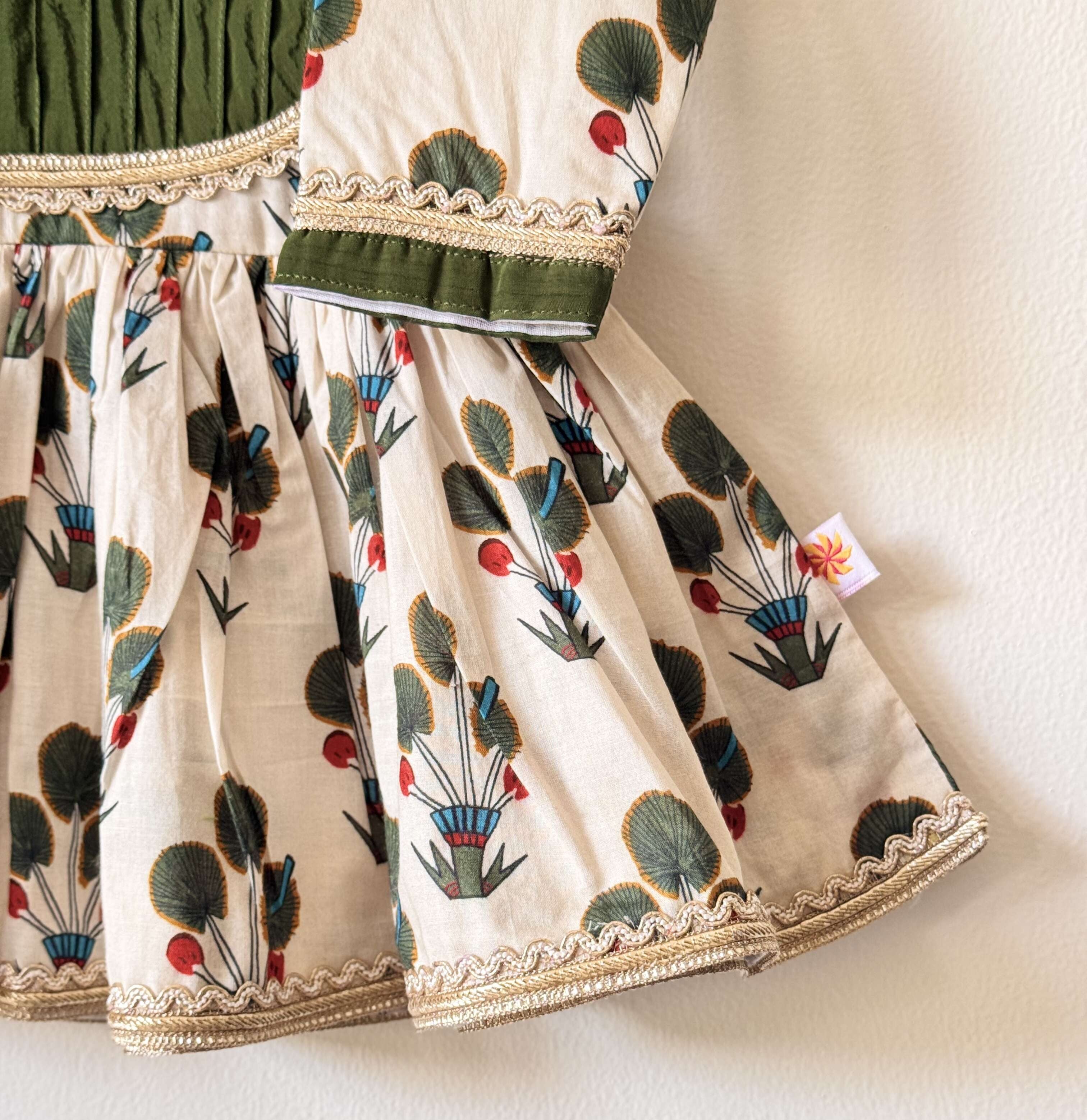 Close-up of an off-white children's flared peplum kurti with green and red floral motifs, featuring a dark green pintuck yoke outlined in gold lace